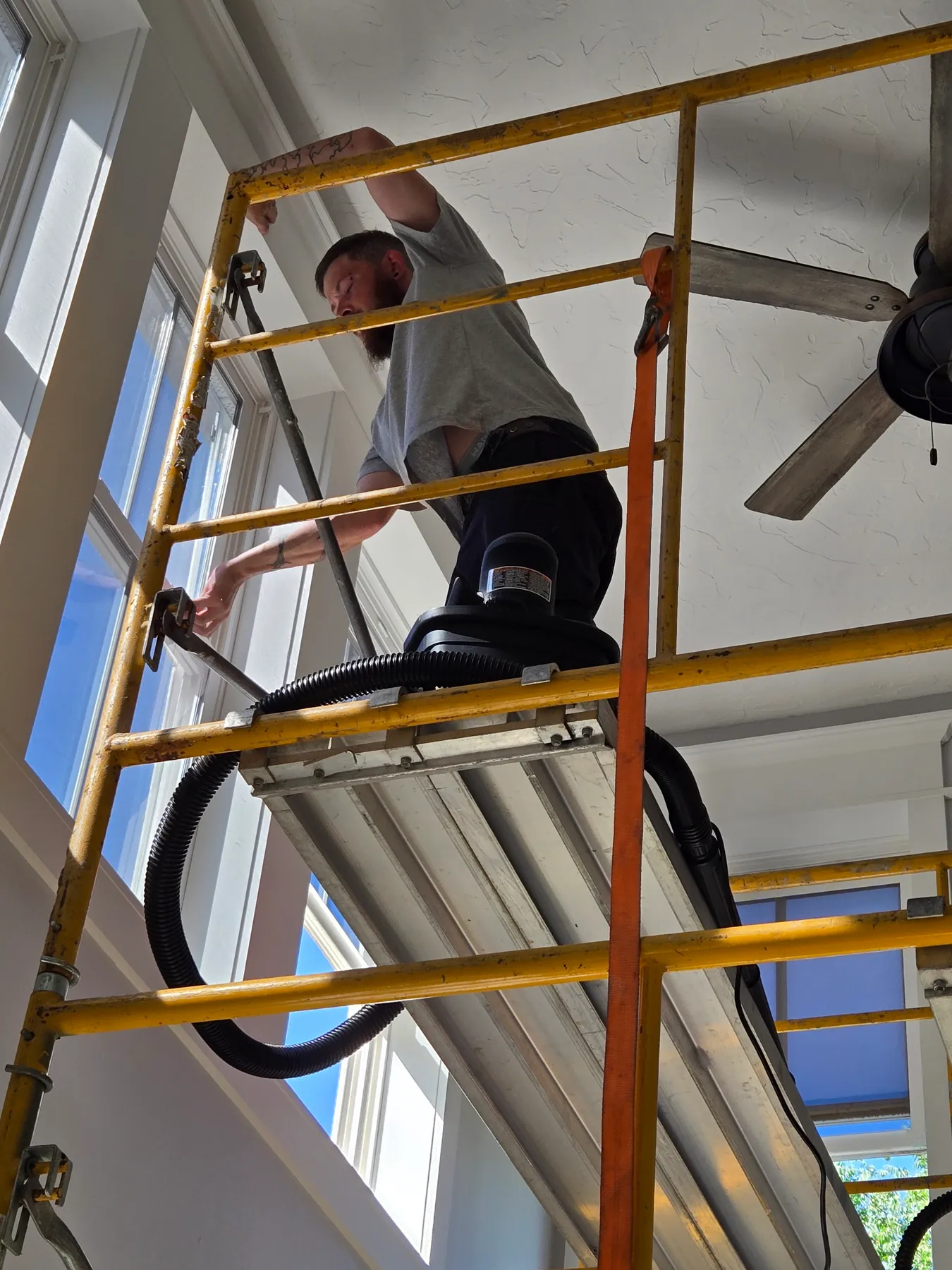Worker on a yellow scaffold beside high interior windows with a vacuum and hose on the platform